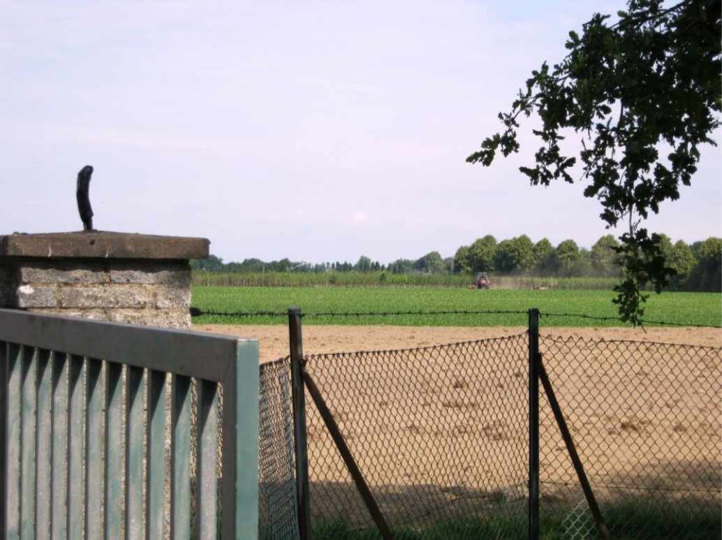 Photograph at the property boundary of the production well field. A wellhead casing in cast iron sits on a concrete pillar in the foreground, behind a metal gate and a chain-link fence with blue mesh. Beyond the fence lies a recently harvested agricultural field with a red tractor visible at distance preparing the soil. A treeline marks the far horizon. Photograph by Osman Can Kandemiroglu.