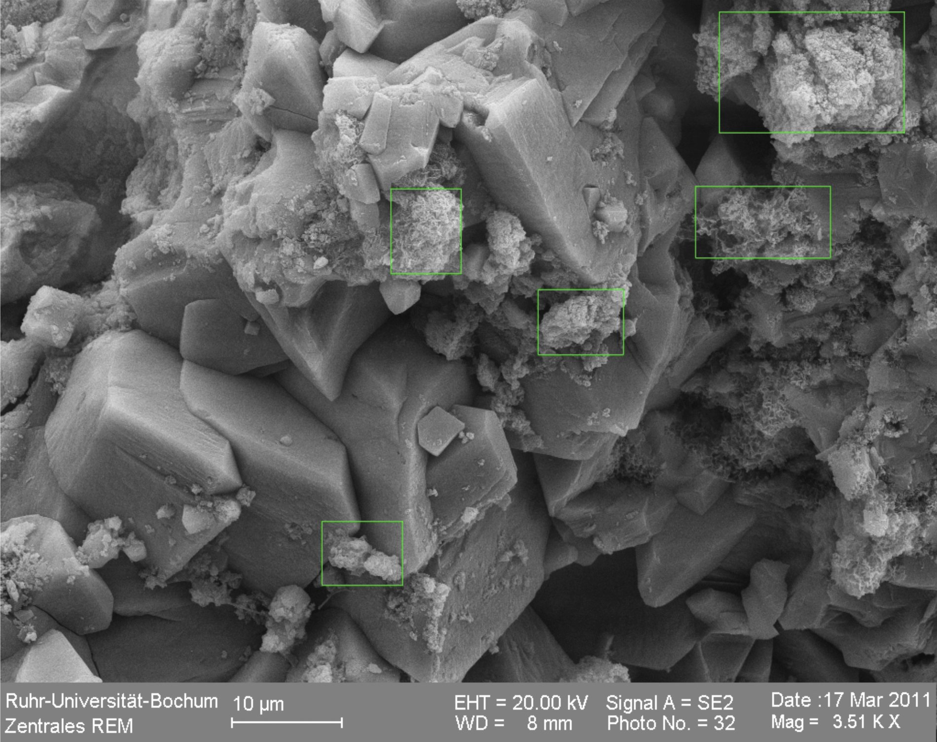 Scanning electron micrograph of spent filter material (sample F2-8): sharp, faceted calcite crystals with iron-oxide coatings marked by green squares; 10 µm scale bar, Ruhr-Universität Bochum.